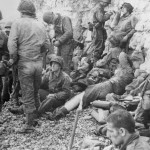A large group of American assault troops of the 3d Battalion, 16th Infantry Regiment, 1st U.S. Infantry Division, having gained the comparative safety offered by the chalk cliff at their backs, takes a breather before moving onto the continent at Colville-Sur-Mer, Omaha Beach, in Normandy, France. Medics who landed with the men treat them for minor injuries.